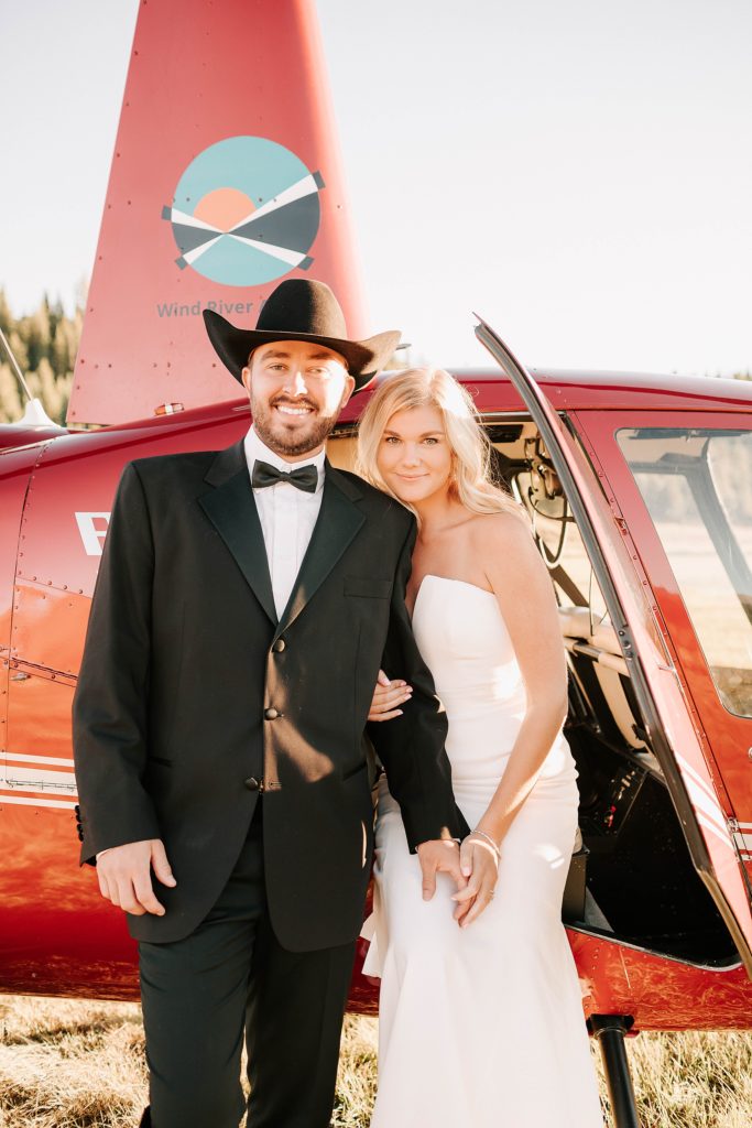 bride and groom standing by red helicopter with the door open, smiling at photographer adrian wayment photo.