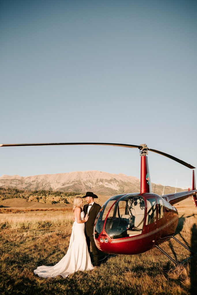 bride and groom facing each other, leaning against a red helicopter near Jackson Hole.