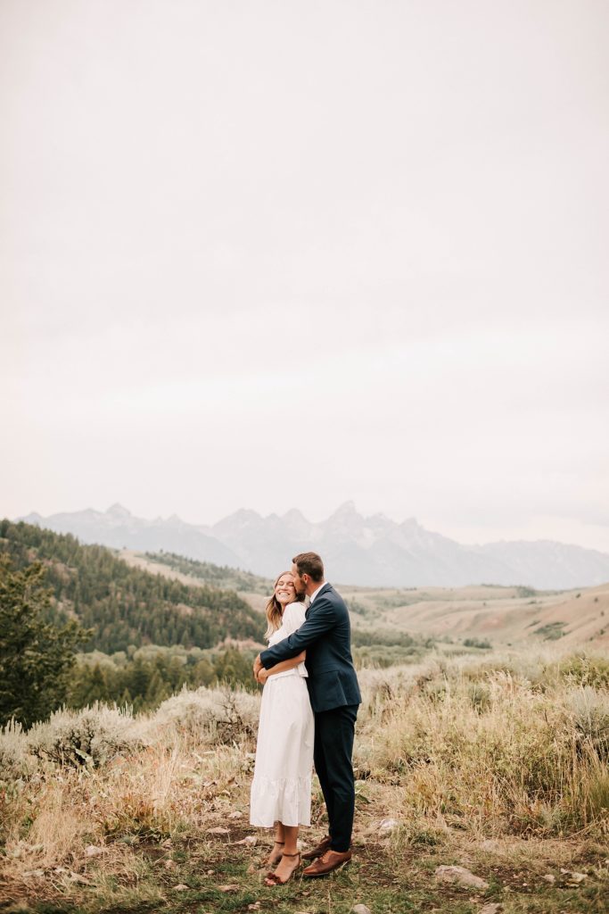 engaged groom hugging bride from the back with the tetons in the background at the wedding tree.