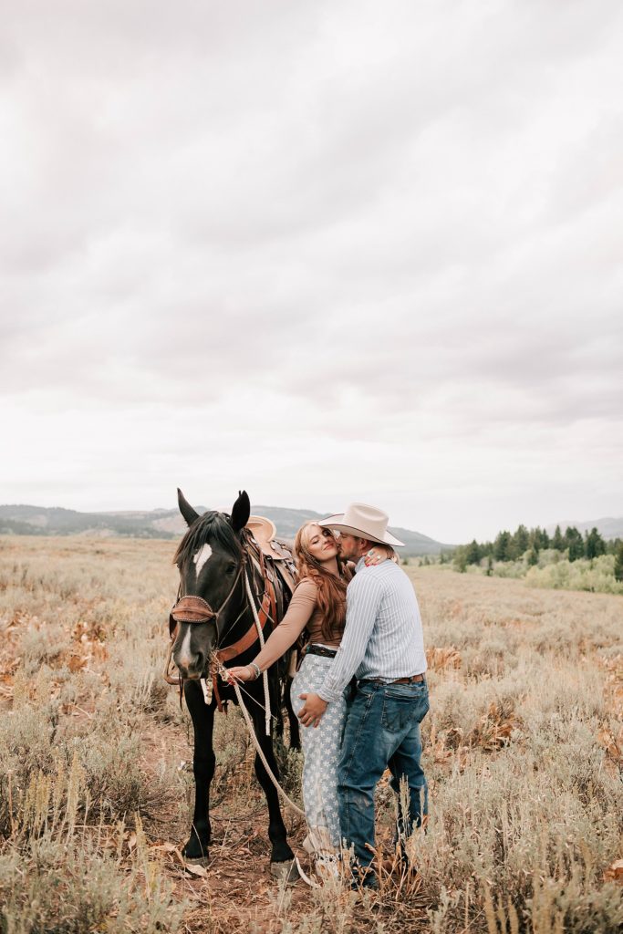 engaged boy and girl standing next to their horse with the guy giving the girl a kiss on the cheek for their western engagement photos.